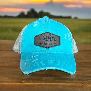 May include: A turquoise blue distressed trucker hat with a black leather patch that says "mother hustler".