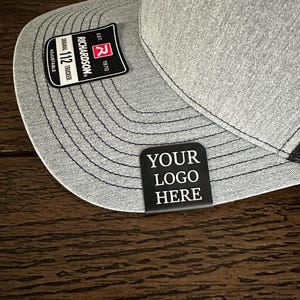 May include: A gray and black trucker hat with a black patch that says "YOUR LOGO HERE" on the front. The hat has a black brim and a gray mesh back. The hat is adjustable and has a label that says "Richardson 112 Original Trucker Est. 1970".