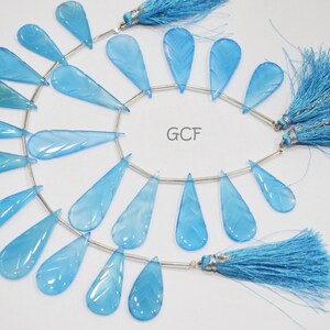May include: A strand of light blue gemstone beads with a carved leaf design. The beads are strung on a silver wire and have a teardrop shape. The strand is finished with two blue tassels.
