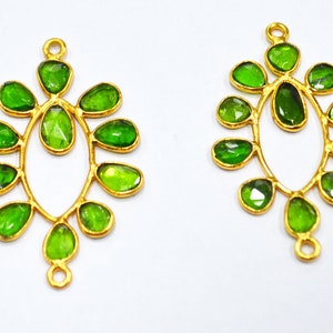 Puede incluir: Dos colgantes de pendientes de metal dorado con detalles de piedras preciosas verdes. Cada colgante presenta una piedra central en forma de lágrima rodeada de piedras más pequeñas de forma ovalada.