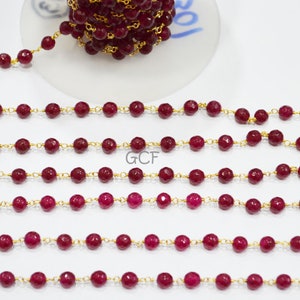 Puede incluir: Una hebra de cuentas de piedras preciosas rojas enhebradas en una cadena de oro. Las cuentas son facetadas y tienen una forma ligeramente irregular. La cadena es delicada y tiene un patrón de eslabones finos. Las cuentas y la cadena están dispuestas en línea recta sobre una superficie blanca.