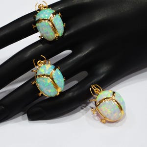 May include: Three gold-coloured beetle pendants with iridescent opal-like bodies. The beetles are displayed on a black surface. The jewellery has a detailed design, with the gold outlining the beetle's shape.