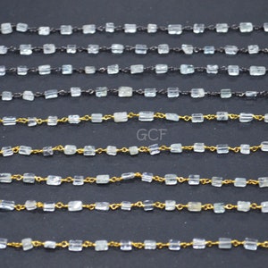 May include: Five strands of gemstone chain, each with a different metal finish. The chains are made of clear faceted beads and gold or black metal links. The chains are laid out horizontally on a dark surface.