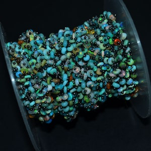 May include: A close-up of a strand of iridescent blue, green, and opal beads with black accents. The beads are strung on a silver chain and are displayed on a clear plastic spool.