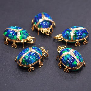 May include: Five gold-coloured beetle pendants with iridescent blue and green enamel bodies. The beetle's legs and details are crafted in gold. The pendants are arranged on a dark surface.