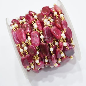 May include: A strand of red gemstone beads with gold chain and white pearl accents. The beads are oval shaped and have a sparkly finish. The strand is wound around a clear spool.