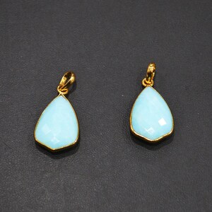 May include: Two teardrop-shaped pendants with a light blue, faceted stone set in a gold-colored metal. The pendants have a loop at the top for a chain. The background is a dark gray color.