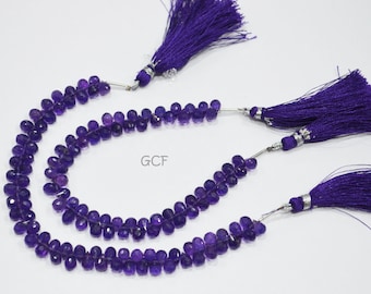 May include: Two strands of purple amethyst beads with faceted surfaces, each strand is strung on a silver wire and finished with a purple tassel. The strands are laid out on a white surface. GCF is printed on the surface.