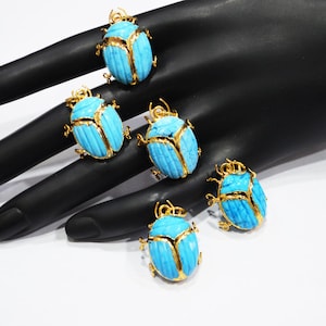 May include: Five turquoise scarab beetle pendants with gold-colored accents are displayed on a black surface. The pendants have a detailed carved design and are arranged to showcase their intricate features. The background is a plain white surface.