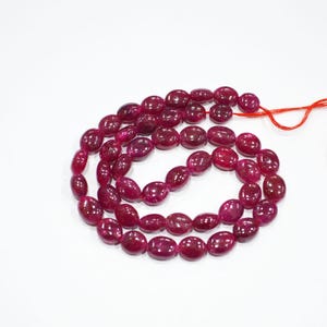 May include: Strand of oval-shaped, deep red gemstones on a white background. The beads are strung together with a red thread, creating a necklace or bracelet. The gemstones have a polished, glossy appearance.
