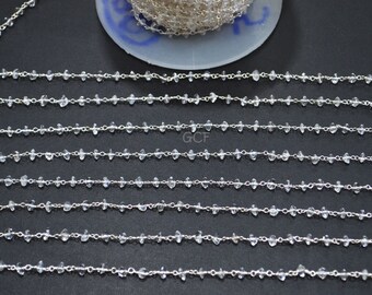 Könnte beinhalten: Nahaufnahme von mehreren Strängen einer Kette aus klarem Quarz-Edelstein. Die Quarzperlen sind unregelmäßig geformt und auf einer Silberkette aufgereiht. Die Ketten liegen auf einer dunklen Oberfläche, im Hintergrund ist eine Kettenspule zu sehen.