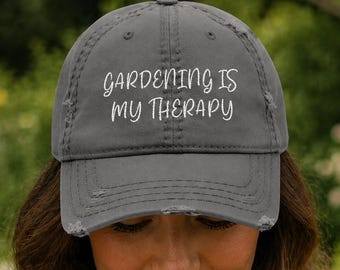 May include: A gray baseball cap with the text "GARDENING IS MY THERAPY" in white script. The cap has a distressed, worn look. The background is a blurred garden scene with green foliage and colorful flowers.