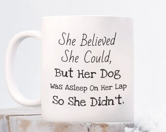 May include: White ceramic coffee mug with black text that reads "She Believed She Could, But Her Dog Was Asleep On Her Lap So She Didn't."