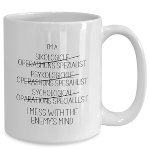 May include: White ceramic coffee mug with black text that reads "I'MA SIKOLOGICLE OPERASHUNS SPEZIALIST PSYKOLOGICKLE OPERASHUNS SPESAHLIST SYCHOLOGICAL OPARATIONS SPECIALLEST I MESS WITH THE ENEMY'S MIND"