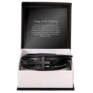 May include: Black leather bracelet with a cross charm, presented in a gift box. The box is black with a white interior. Text inside the box reads "Happy 60th Birthday!" and a message.