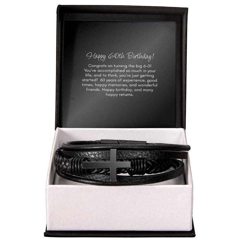 May include: Black leather bracelet with a cross charm, presented in a gift box. The box is black with a white interior. Text inside the box reads "Happy 60th Birthday!" and a message.