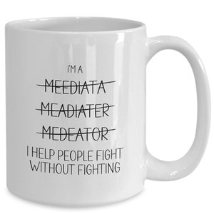 May include: White ceramic coffee mug with black text that reads "I'm a Meeediata Mead이터 Medebtor I help people fight without fighting."