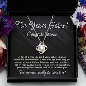 May include: A black gift box containing a silver necklace. The necklace features a knot design with a clear crystal at its centre. The box displays a message that reads "Five Years Sober! Congratulations" and "A day at a time you are 5 years sober, what an incredible achievement! It hasn't always been easy but no matter what life has thrown at you, you handled it sober. Happy, joyous and free, you are an inspiration, your strength is immense and I'm so proud of you. The promises really do come true!"
