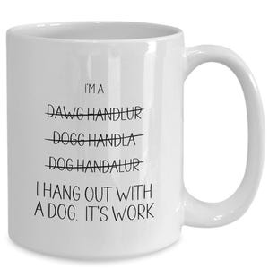 May include: White ceramic coffee mug with black text that reads "I'MA DAWG HANDLUR DOGG HANDLA DOG HANDALUR I HANG OUT WITH A DOG. IT'S WORK"