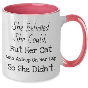May include: White ceramic coffee mug with a pink rim and handle. The mug has black text that reads "She Believed She Could, But Her Cat Was Asleep On Her Lap So She Didn't."