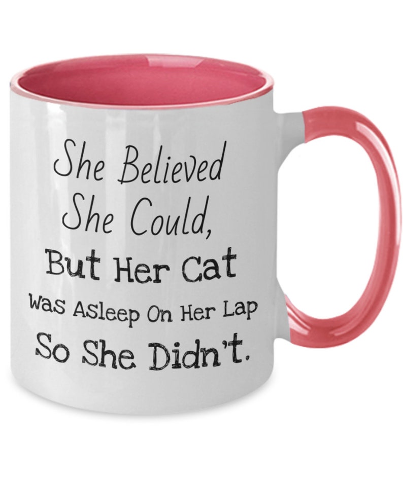 May include: White ceramic coffee mug with a pink rim and handle. The mug has black text that reads "She Believed She Could, But Her Cat Was Asleep On Her Lap So She Didn't."