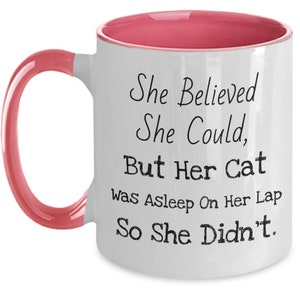 May include: White ceramic coffee mug with a pink rim and handle. The mug has black text that reads "She Believed She Could, But Her Cat Was Asleep On Her Lap So She Didn't."