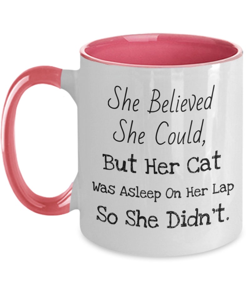 May include: White ceramic coffee mug with a pink rim and handle. The mug has black text that reads "She Believed She Could, But Her Cat Was Asleep On Her Lap So She Didn't."