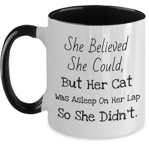 May include: White ceramic coffee mug with a black rim and handle. The mug has a funny saying printed on it: "She Believed She Could, But Her Cat Was Asleep On Her Lap So She Didn't."