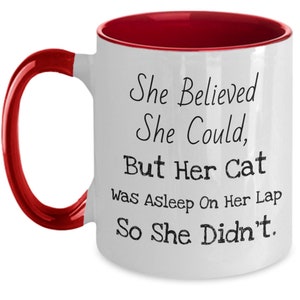 May include: White ceramic coffee mug with a red rim and handle. The mug has a funny saying printed on it: "She Believed She Could, But Her Cat Was Asleep On Her Lap So She Didn't."