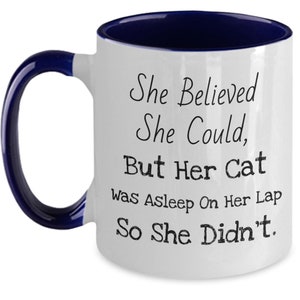May include: White ceramic coffee mug with a blue rim and handle. The mug has a funny saying printed on it: "She Believed She Could, But Her Cat Was Asleep On Her Lap So She Didn't."
