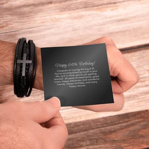 May include: A black leather bracelet with a silver cross charm is shown next to a black card. The card reads "Happy 60th Birthday!" with a message congratulating the recipient on their milestone.