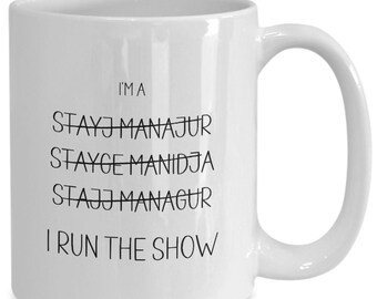 May include: White ceramic coffee mug with black text that reads "I'm a Stay-at-Home Manager, Stay-at-Home Manidja, Stay-at-Home Manager, I Run The Show."