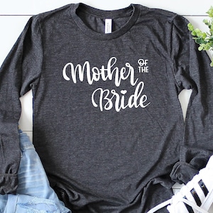 May include: A dark gray long-sleeved shirt with the text "Mother of the Bride" in white lettering. The shirt has a relaxed fit and is perfect for a wedding.