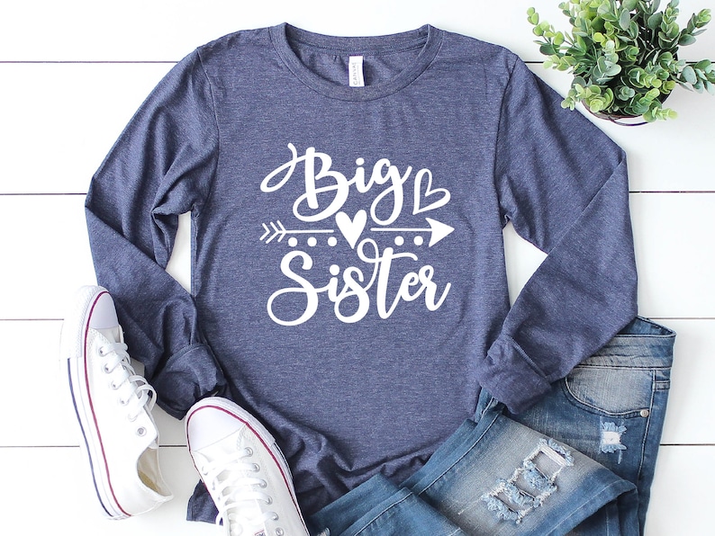 May include: A navy blue long-sleeved shirt with the text "Big Sister" in white lettering. The text includes a heart and an arrow.