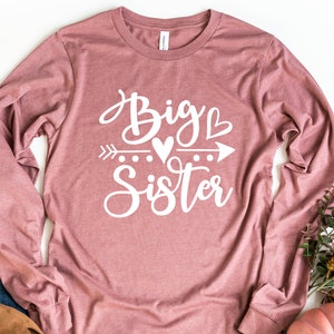 May include: A long-sleeved shirt in a dusty rose color with the text "Big Sister" in white lettering with a heart and arrow graphic.