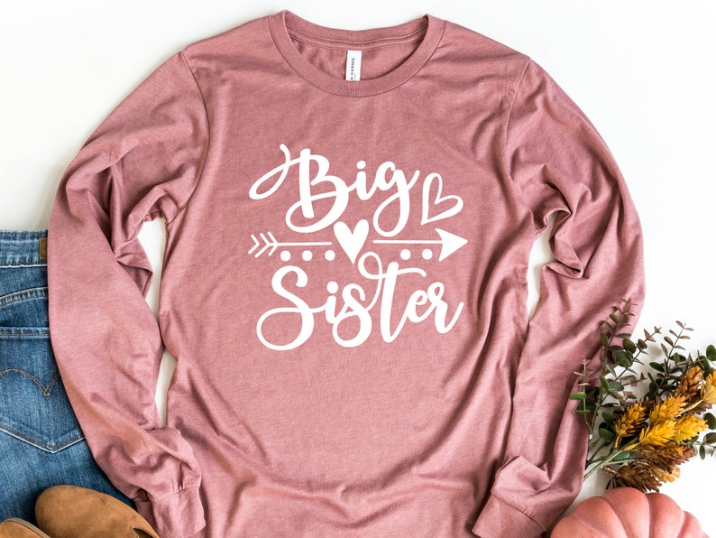 May include: A long-sleeved shirt in a dusty rose color with the text "Big Sister" in white lettering with a heart and arrow graphic.