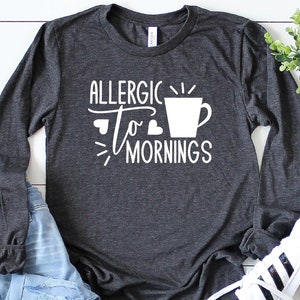May include: A dark gray long-sleeved shirt with the text "Allergic to Mornings" in white, featuring a stylized coffee cup graphic.