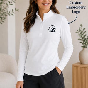May include: A white long-sleeve quarter-zip pullover featuring a black embroidered mountain and pine tree logo on the chest, paired with black athletic shorts. The text Custom Embroidery Logo is visible with an arrow pointing to the chest.