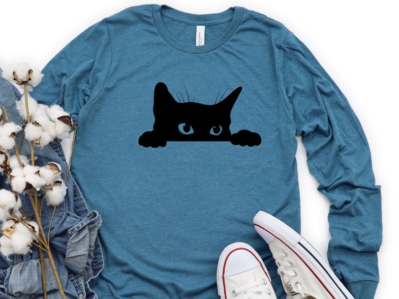 May include: A teal blue long-sleeved t-shirt featuring a black silhouette of a cat peeking over the top. The cat has a white eye and is looking directly at the viewer.