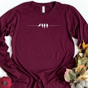 May include: A maroon long-sleeved t-shirt with a white silhouette of four birds perched on a wire.