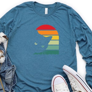 May include: A teal blue long-sleeved t-shirt with a retro sunset graphic featuring a cat silhouette. The cat is peeking out from behind the sunset, which has a rainbow of colors.