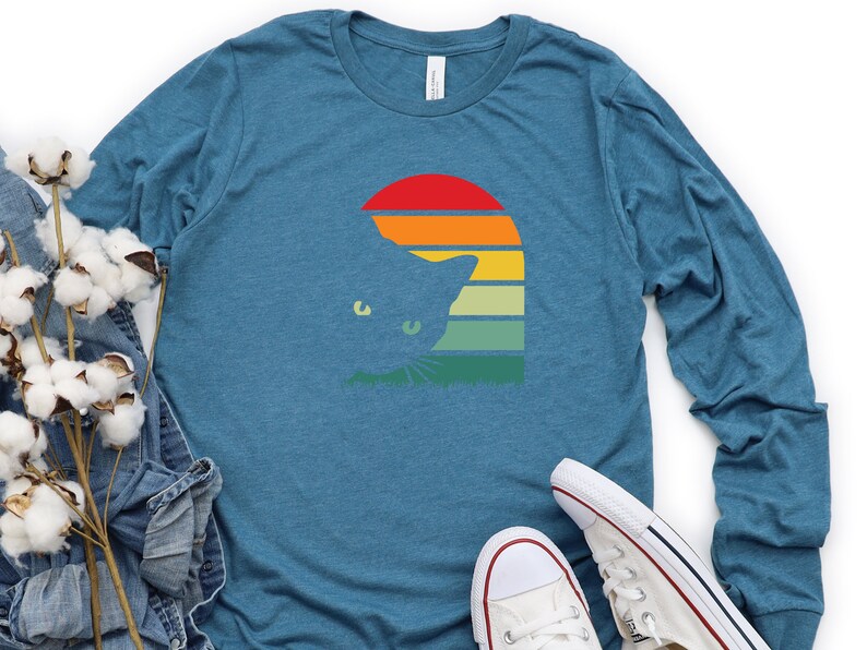 May include: A teal blue long-sleeved t-shirt with a retro sunset graphic featuring a cat silhouette. The cat is peeking out from behind the sunset, which has a rainbow of colors.
