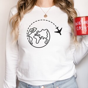 May include: White long-sleeve shirt with a black outline of a globe and airplane, suggesting travel. The shirt is worn by a person holding a red mug with the text "warm cozy" in white.