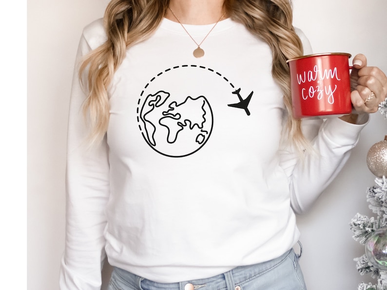 May include: White long-sleeve shirt with a black outline of a globe and airplane, suggesting travel. The shirt is worn by a person holding a red mug with the text "warm cozy" in white.