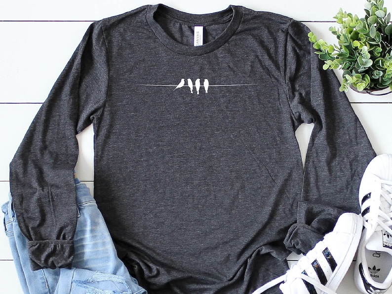 May include: A dark gray long-sleeved t-shirt with a white graphic of five birds on a wire. The shirt is made of a soft, comfortable material.