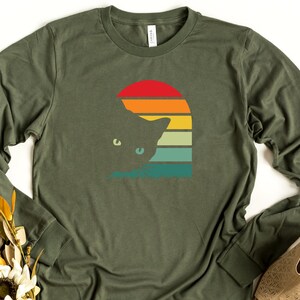 May include: A green long-sleeved t-shirt with a retro sunset graphic featuring a cat silhouette. The sunset is in shades of orange, yellow, green, and blue.