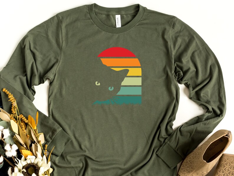 May include: A green long-sleeved t-shirt with a retro sunset graphic featuring a cat silhouette. The sunset is in shades of orange, yellow, green, and blue.