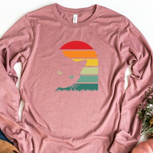 May include: A long-sleeved, mauve t-shirt with a retro-style graphic of a cat peeking out from behind a sunset. The graphic features a red, orange, yellow, green, and blue color scheme.