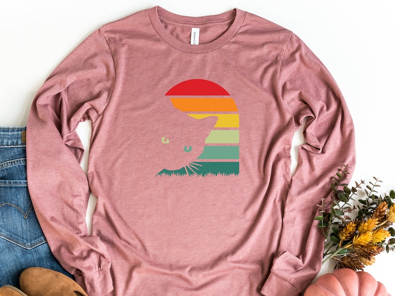 May include: A long-sleeved, mauve t-shirt with a retro-style graphic of a cat peeking out from behind a sunset. The graphic features a red, orange, yellow, green, and blue color scheme.
