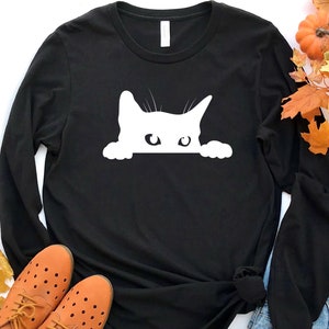 May include: Black long-sleeved t-shirt with a white silhouette of a cat peeking out from behind the fabric.
